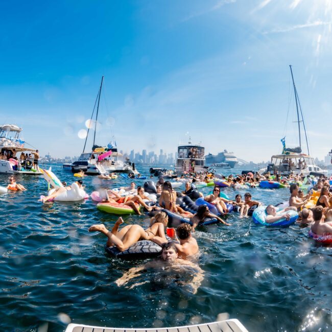 A vibrant scene of people enjoying a sunny day on the water. Dozens of people are on colorful inflatables or swimming, surrounded by boats. The city skyline is visible in the background under a clear blue sky.