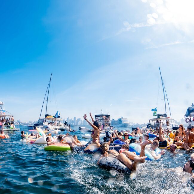 A lively scene on the water with people lounging on inflatable rafts and floating in swimsuits. Boats and yachts are in the background under a clear blue sky, and city skyscrapers are faintly visible in the distance.
