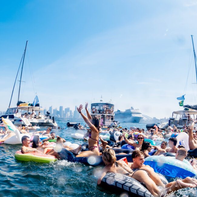 A large group of people floats on various inflatable toys in a sunny harbor, with sailboats and a cruise ship in the background. The skyline of a city is visible under a clear blue sky.