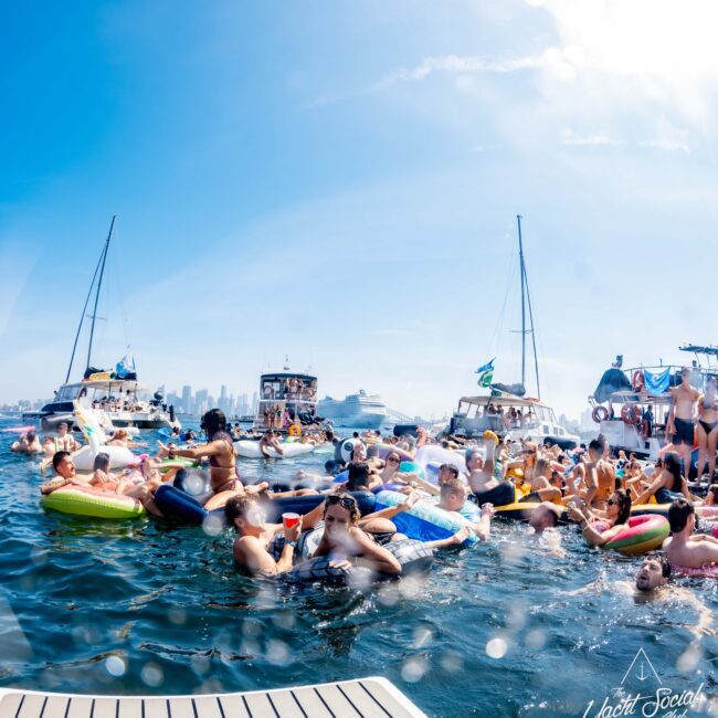 A lively scene of people enjoying a sunny day on the water with boats and inflatable floats. The background features a city skyline. A logo in the corner reads "Yacht Social Club.
