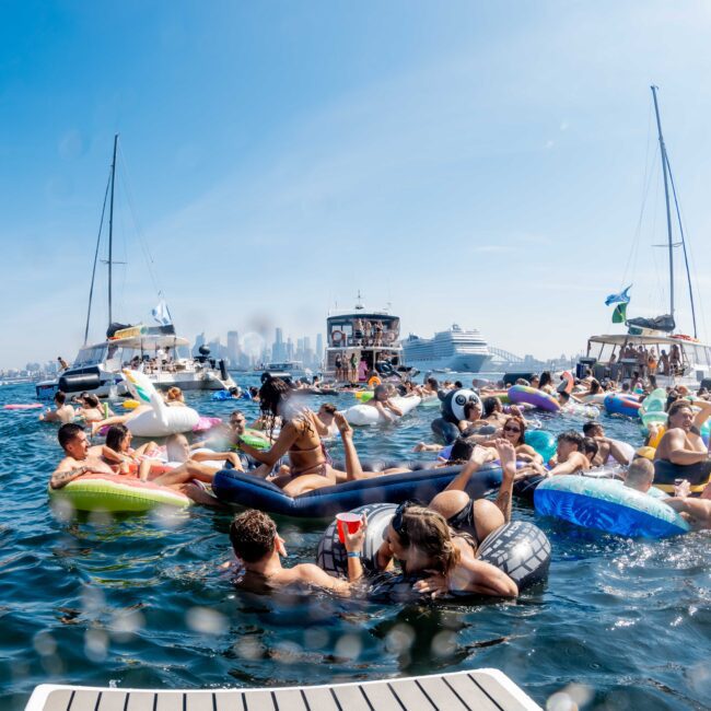 People gather on inflatable rafts and floaties in a lively, crowded marina, surrounded by several boats. The background shows a city skyline. The atmosphere is festive, with sunny weather and clear blue skies.