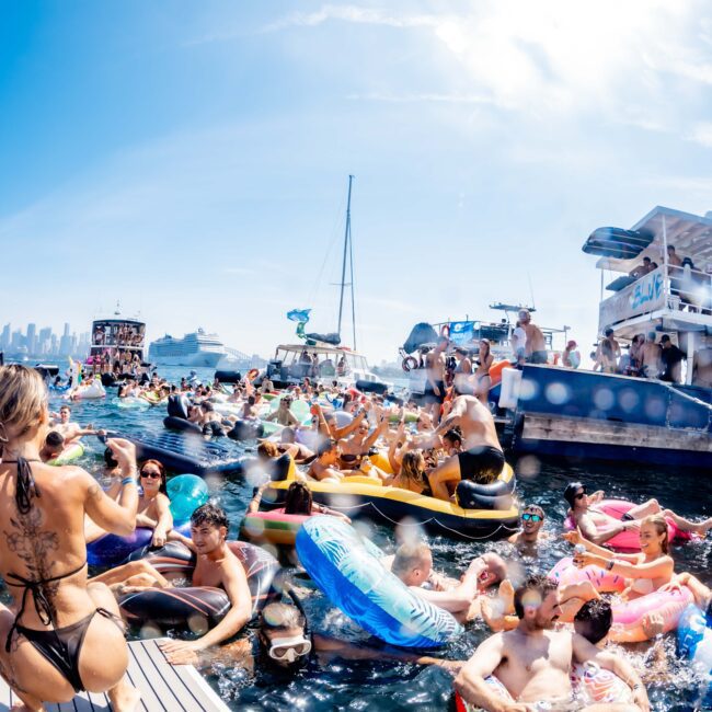 A lively boat party with people enjoying the water on colorful inflatables. Boats are anchored nearby with music and dancing. The sky is clear, and the city skyline is visible in the background.