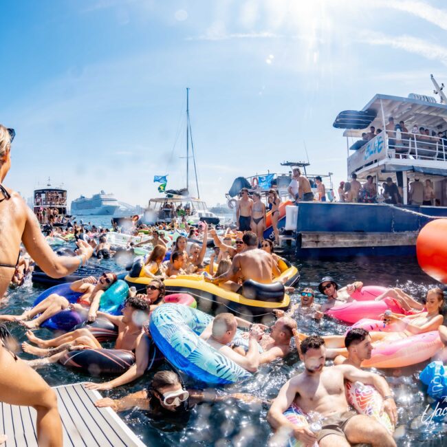 A crowded scene with people in swimwear enjoying a sunny day on a boat party. Many are floating on colorful inflatable devices in the water, while others are gathered on the deck of a nearby yacht. The atmosphere is lively and festive.