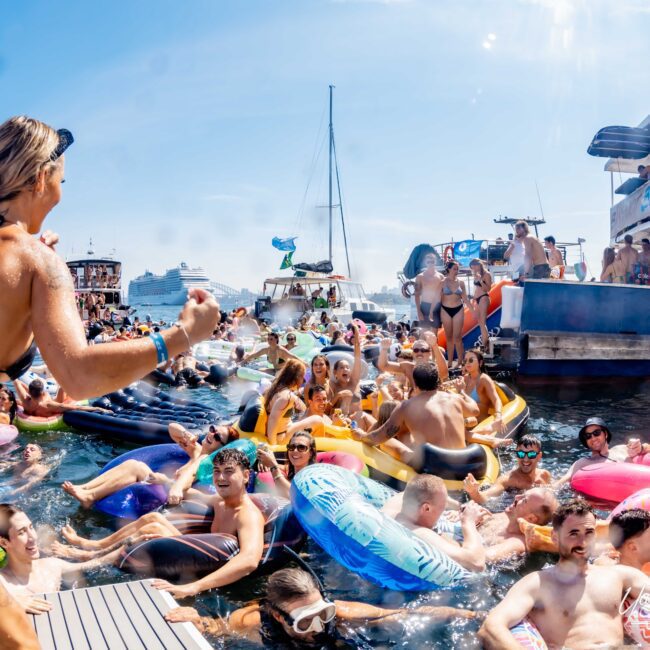 A crowded water party scene with people in swimsuits on inflatable floats near a dock. Boats are visible in the background on a sunny day. Some attendees are in the water while others dance on the platform. The mood is lively and festive.