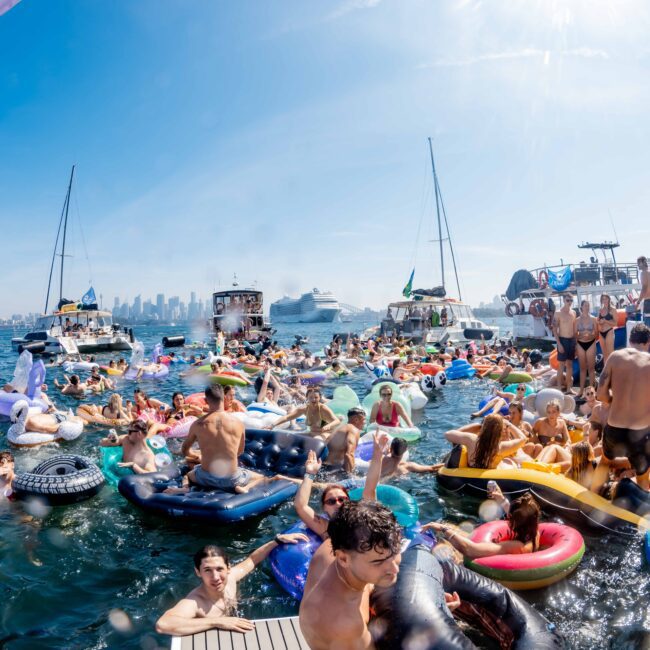 A crowded scene of people enjoying a sunny day on water inflatables and boats. Various colorful floats are visible, with a cityscape and cruise ship in the background. The atmosphere is lively and festive.