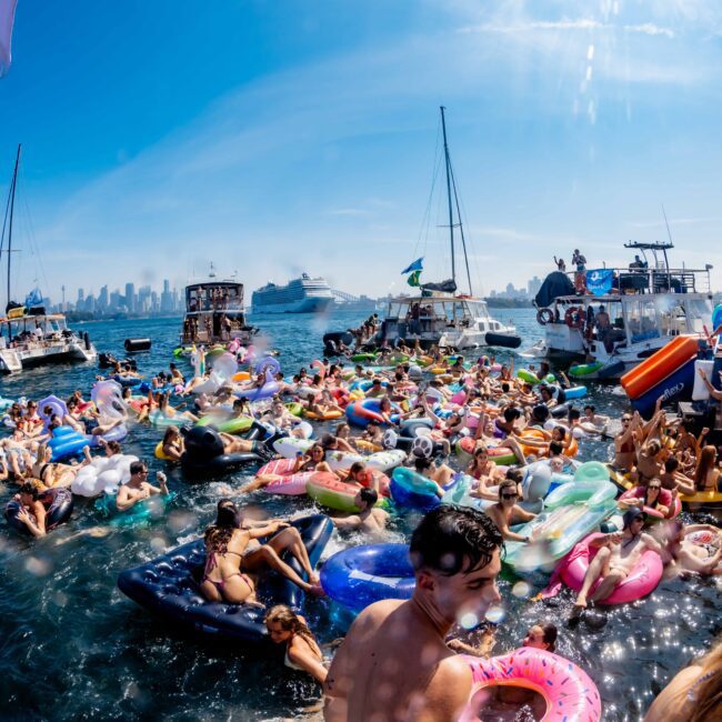 A lively scene of a crowded boat party near a city skyline. People are enjoying the water on colorful inflatables, surrounded by several yachts and boats. The atmosphere is festive under a clear blue sky.