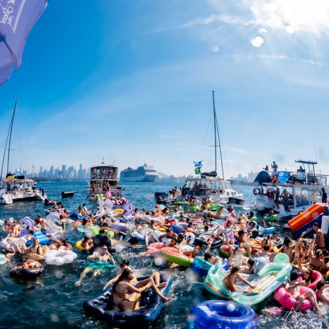 Crowd of people in a marina enjoying a sunny day on colorful inflatables. Several boats surround the area, with a city skyline visible in the background. The atmosphere is festive and lively.