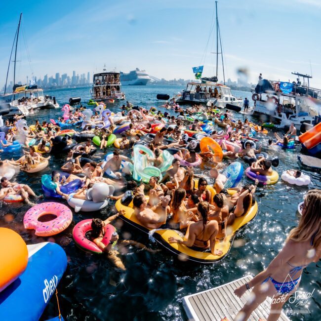 A crowded pool party scene with numerous people on colorful inflatable floats, including flamingos and donuts, in a body of water. Boats surround the area, and a city skyline is visible in the background under a clear blue sky.