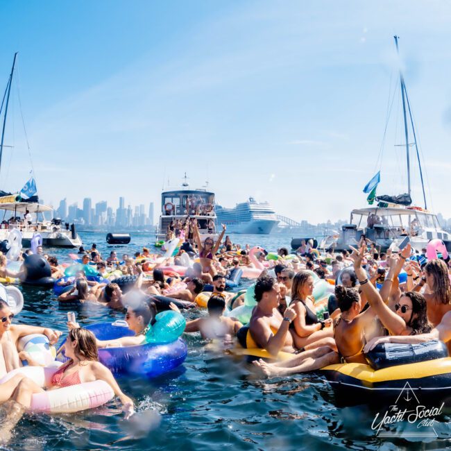 A lively scene of people on inflatable rafts and tubes in the water, celebrating near yachts and a large cruise ship. The skyline of a city can be seen in the background under a clear blue sky.
