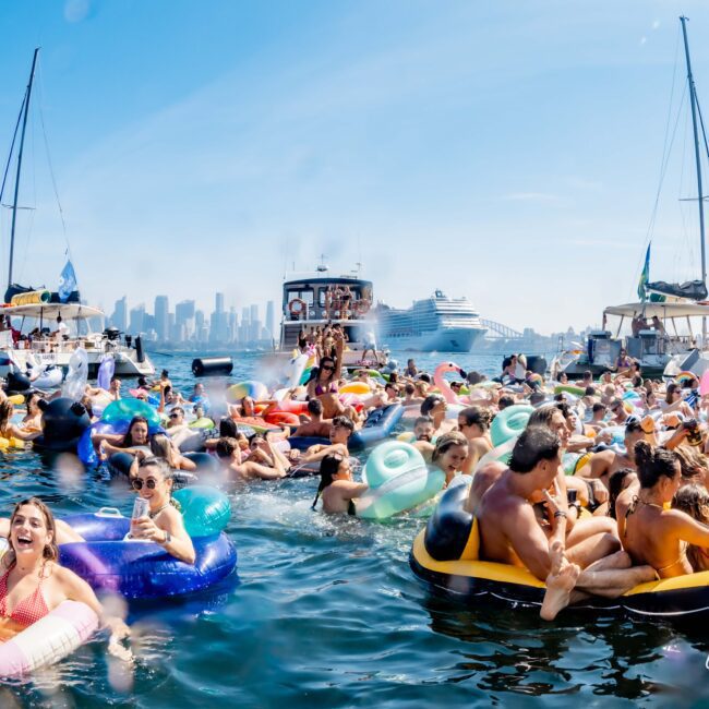 A lively scene on the water shows a large group of people enjoying a party while floating on inflatable floats. Sailboats and a cruise ship are in the background, with a city skyline visible under a clear blue sky.