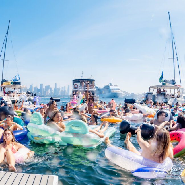 A crowded beach party with people floating on colorful inflatables in the water. Boats and a cruise ship are visible in the background with a city skyline under a clear blue sky.