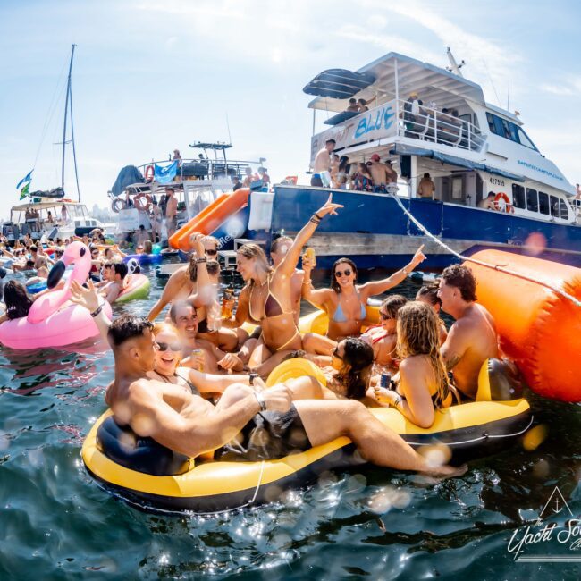 A lively scene showing a group of people in swimsuits enjoying a party on inflatable rafts in the water. A boat named "Blue" is in the background. There's a playful atmosphere with floats like a pink flamingo, under a sunny sky.