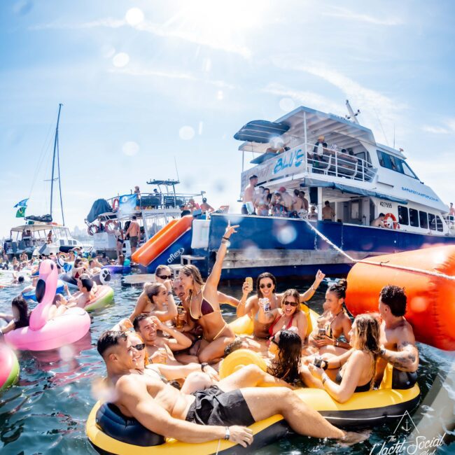 A group of people enjoy a sunny day on a lake, lounging on inflatable rafts. They are socializing and holding drinks near a large boat named "Blue." Other boats and inflatables are visible in the background.