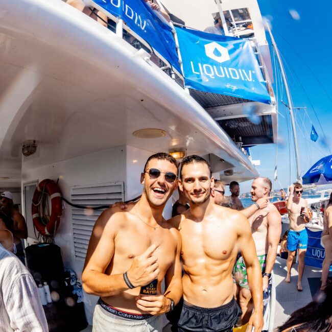 Two men pose and smile while standing on a boat deck. They are shirtless and wearing shorts. Behind them, people socialize under a blue LiquiD banner. The sky is clear and sunny.
