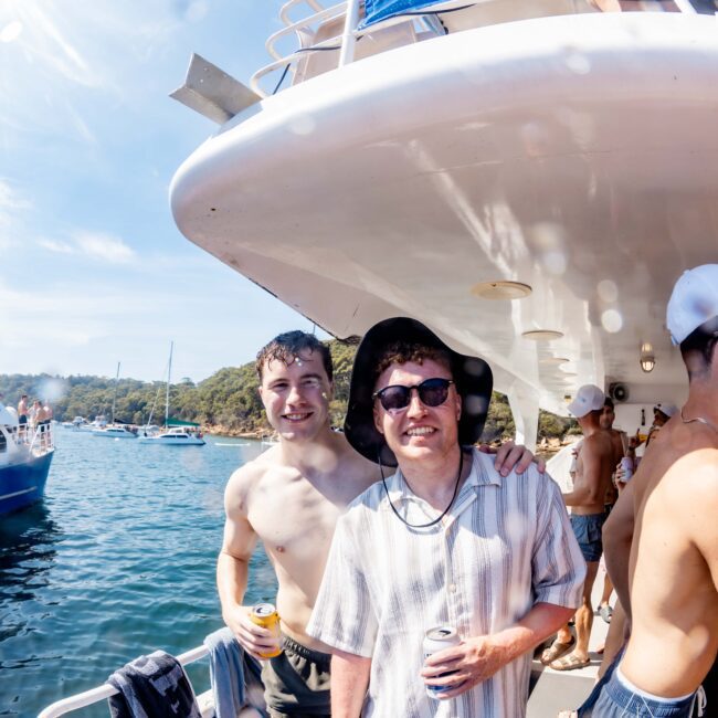 Two people stand on a boat deck, both holding drinks. One is shirtless; the other wears a striped shirt and sunglasses. They're smiling and posing for the camera. Other boats and people are visible in the background.