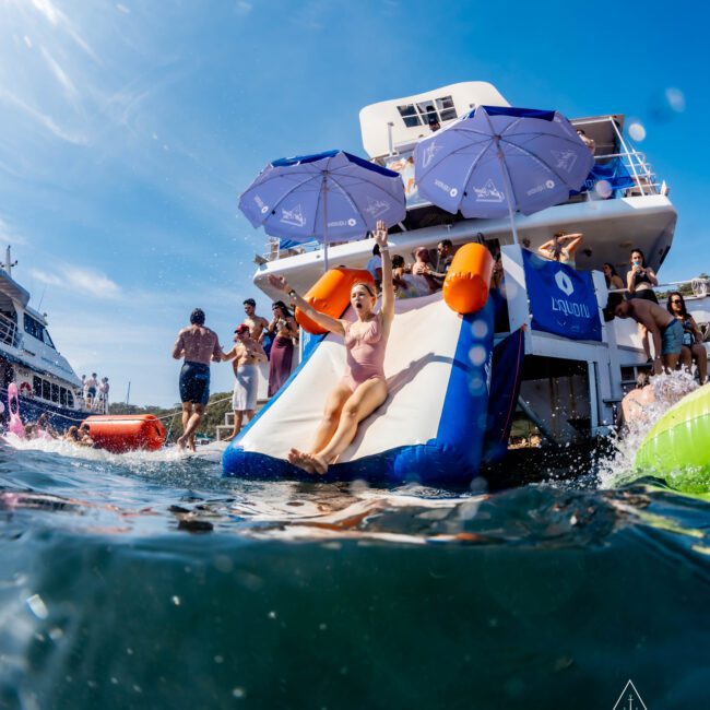 A person slides down an inflatable slide into the ocean from a yacht with blue umbrellas and people watching. Other boats and people are visible in the background under a clear blue sky.