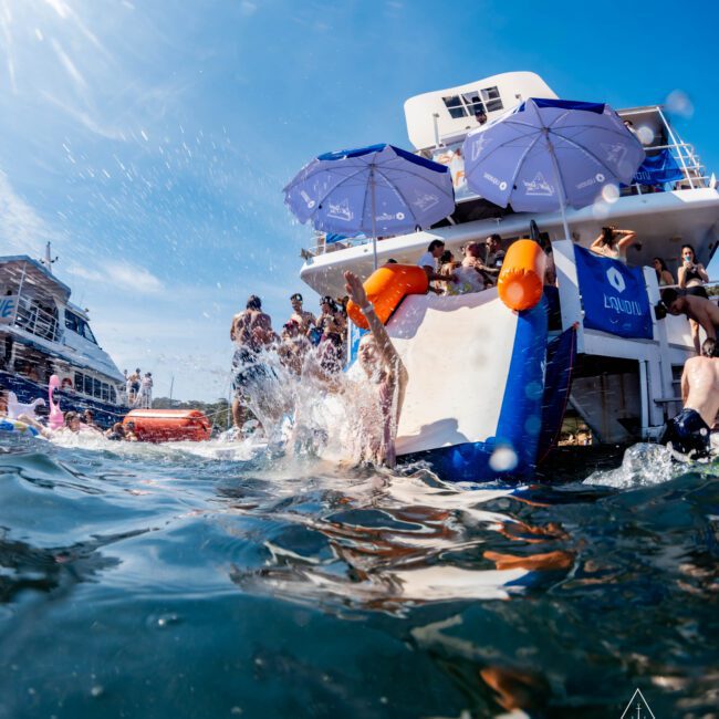People enjoying a sunny day on a boat with blue umbrellas. A person is sliding into the water while others relax nearby. The boat features a "LayOn" sign. Splashes and vibrant activities suggest a lively and fun atmosphere.