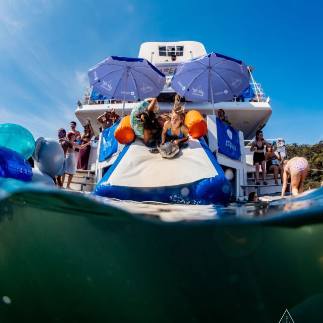 People are enjoying a sunny day on a yacht with a water slide. Some are in swimsuits, descending the slide into the water, while others watch from the deck. Blue umbrellas are open, and there's a mix of excitement and relaxation.