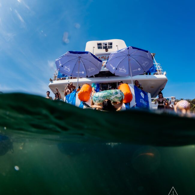 A split-shot image showing a party on a boat above water, with people and blue umbrellas, and a swimmer wearing orange arm floaties underwater. The boat has a logo with "The Yacht Social Club" visible on the bottom right.