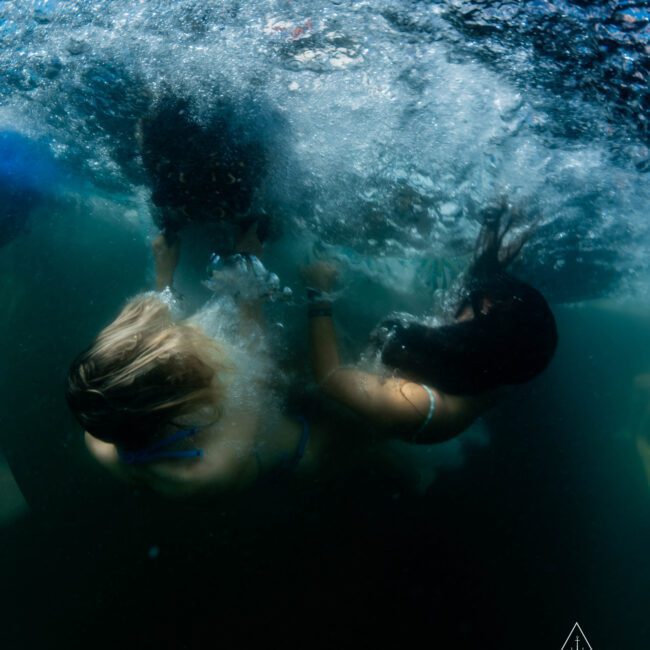 Two people underwater in a bubbly, greenish environment. They are partially submerged, with one facing down and the other facing up. The scene is dynamic, with water movement and splashes visible around them.