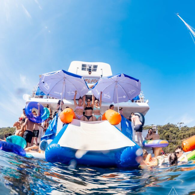 People are enjoying a sunny day on a boat with large umbrellas. Some are lounging on an inflatable slide leading into blue water. The backdrop features a clear sky and wooded hills.