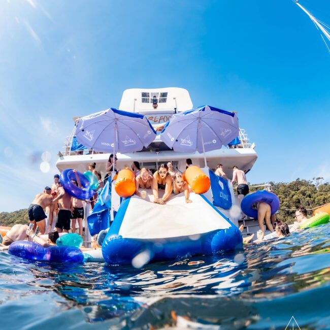 A group of people enjoys a sunny day on a yacht equipped with a slide. Some are on inflatable rafts in the water, while others are on the slide holding inflatable floats. Trees and clear skies are in the background.