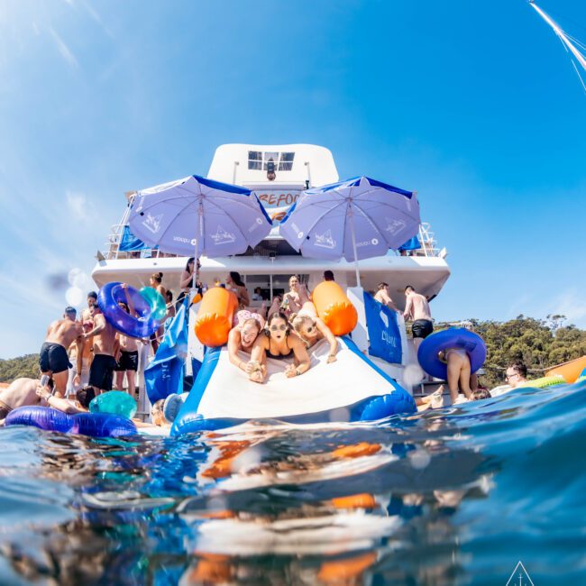 People enjoying a sunny day on a yacht with umbrellas and a large inflatable slide into the water. Swimmers with floats are in the foreground. The sky is clear and blue.