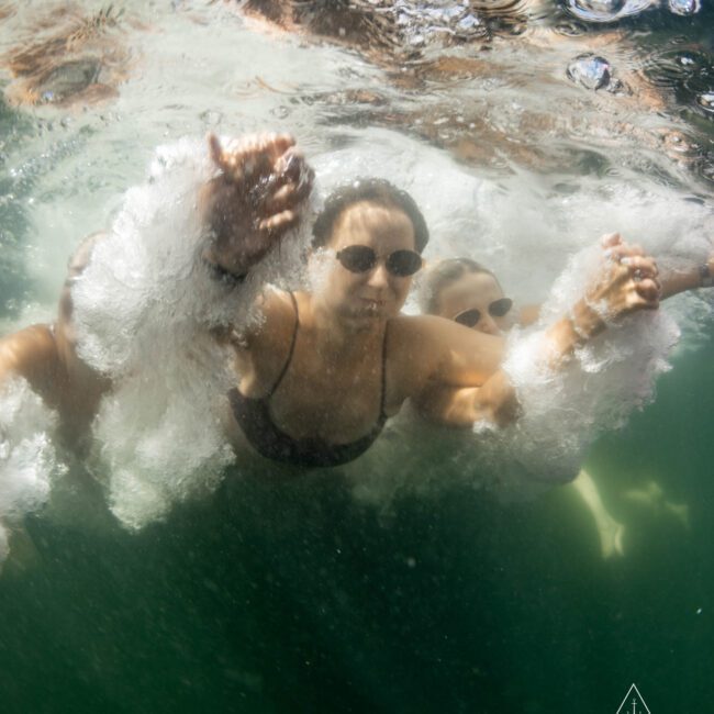 A group of people underwater, wearing swimsuits and sunglasses, creating bubbles as they swim toward the surface. The lighting highlights the water's movement. A logo in the corner reads "The Yacht Social Club.