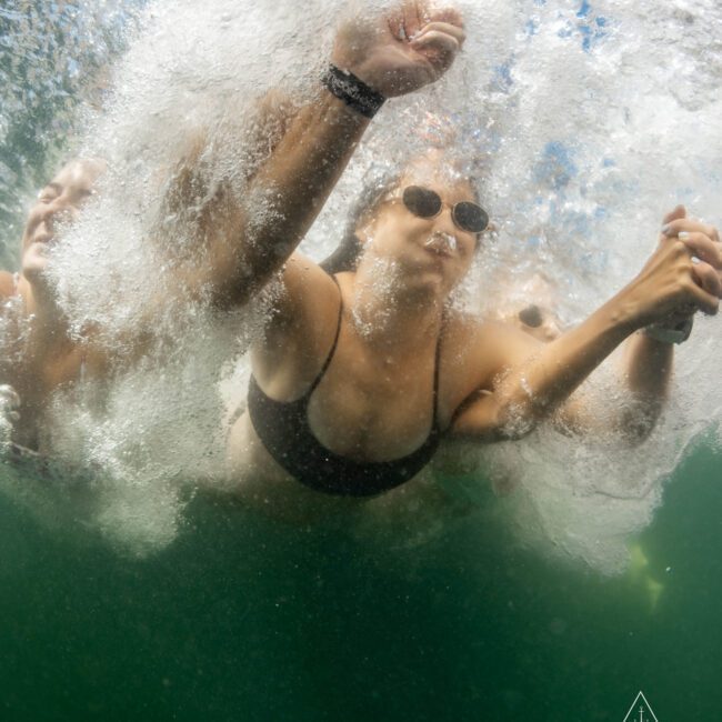 A person wearing sunglasses and a black swimsuit is underwater, surrounded by bubbles and splashes. They are holding hands with another person, creating a dynamic scene of movement beneath the water's surface.