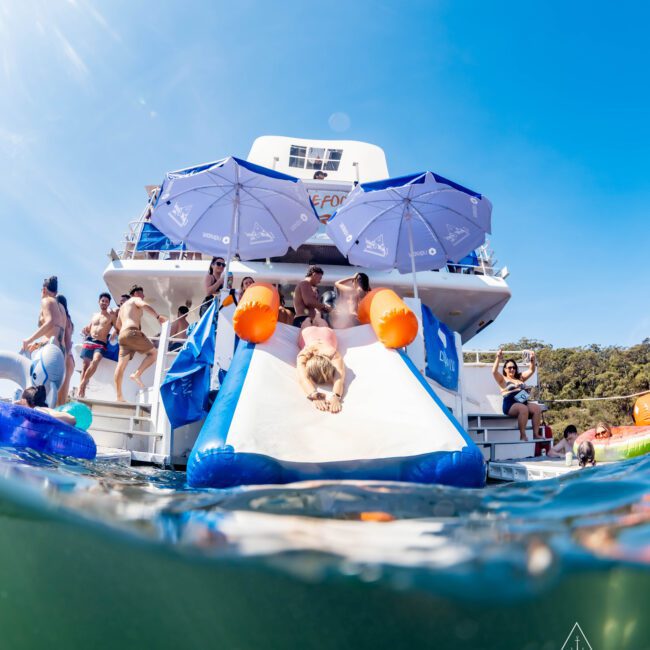 People enjoying a sunny day on a yacht with a slide leading into the water. Blue sky and trees in the background, with party-goers relaxing under umbrellas and swimming in the sea. A person is sliding down into the water.