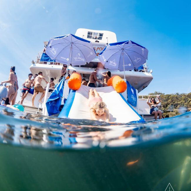 A child slides down an inflatable slide into the water from a boat. Adults relax on the boat, some under blue umbrellas. The image is split between above and below the waterline, capturing a fun, sunny scene.