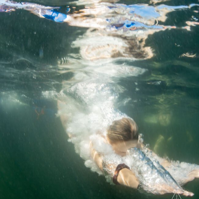 A person swimming underwater in a clear body of water, creating bubbles around them. The water surface above reflects the scene below. There is a logo in the lower right corner that reads "The Yacht Social Club.