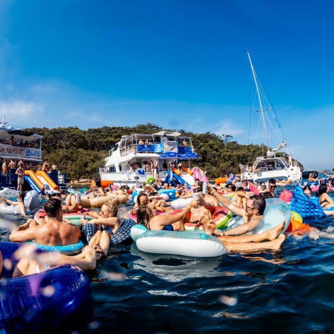 A lively crowd of people relax and enjoy themselves on colorful inflatable floats in a sunny body of water. Yachts and sailboats are moored nearby, while a forested island is visible in the background under a clear blue sky.
