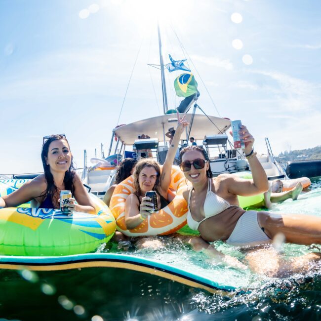 Three people lounge on inflatable rafts in the water, holding drinks. Behind them, a yacht with a Brazilian flag and other boats are visible under a clear blue sky. The atmosphere is festive and relaxed.