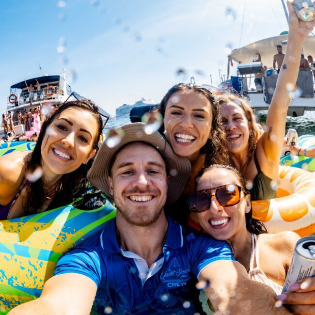 A group of five people smiles at the camera while floating on colorful inflatable rings in the water. They appear to be enjoying a sunny day, with boats and other people visible in the background. One person holds a drink can.