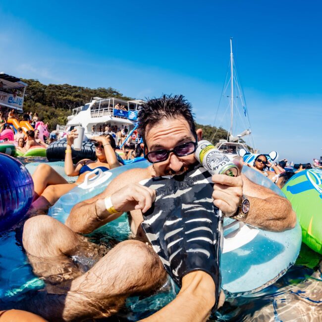 A man floats in the water wearing sunglasses, playfully biting a flipper on another person's foot. He's surrounded by people on colorful inflatables. A boat and sailboat are visible in the background under a clear blue sky.