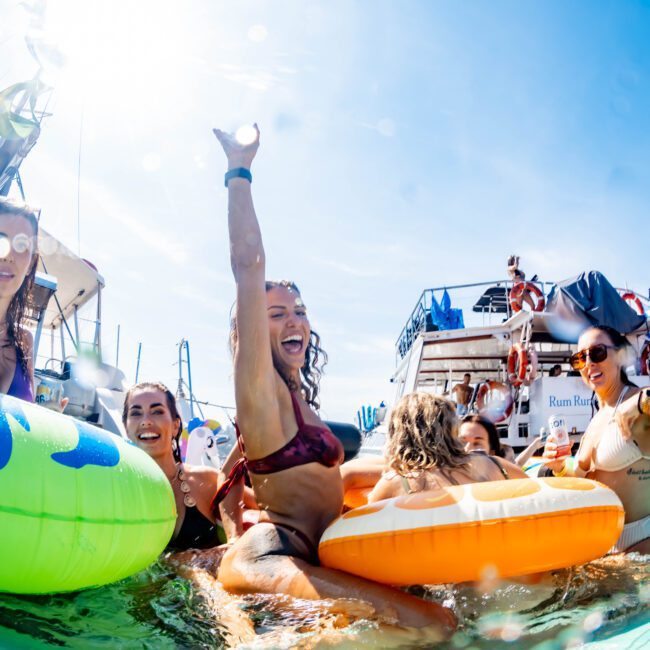 Group of people enjoying a sunny day in the water, surrounded by colorful inflatable floats. A woman in a bikini is raising her arm and smiling, while several boats are visible in the background. The atmosphere is lively and festive.