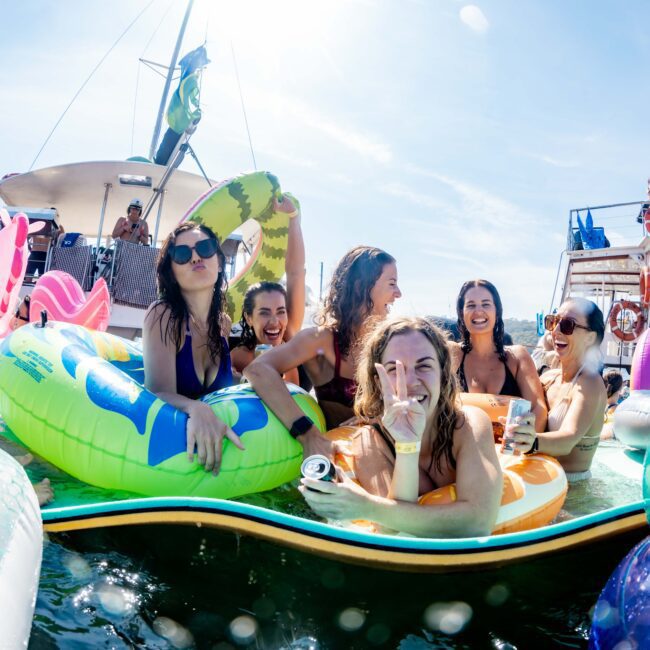 A group of people smiling and having fun in a pool with inflatable floats. They are near a boat under a clear blue sky. The atmosphere is lively and relaxed.
