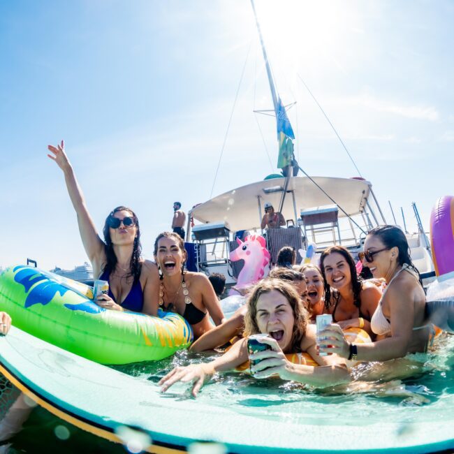 A group of people having fun in the water near a boat. They are sitting on inflatable floats and holding drinks, with one person raising an arm in excitement. The background shows a catamaran and clear blue sky.