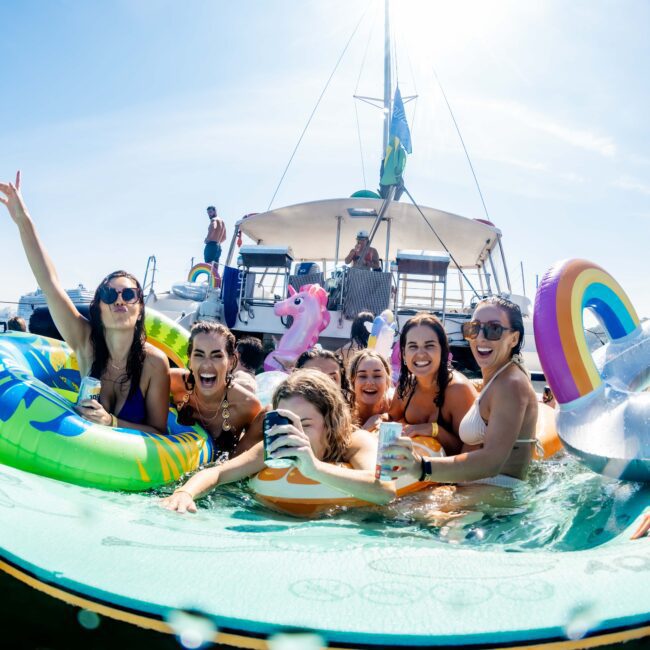 A group of people enjoying a sunny day on the water, surrounded by floats and inflatables near boats. They are smiling, holding drinks, and sitting on a large floating platform.