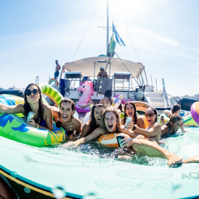A group of people enjoying a party on a boat with colorful inflatables and a unicorn float in the water. They are smiling and having fun while the sun shines brightly. A city skyline is visible in the background.