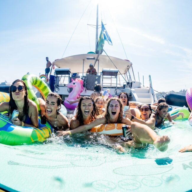 A group of people enjoying a sunny day on the water with inflatable rafts near a boat. The scene is lively, with everyone smiling and laughing. A unicorn float and other inflatables add a playful vibe to the gathering.