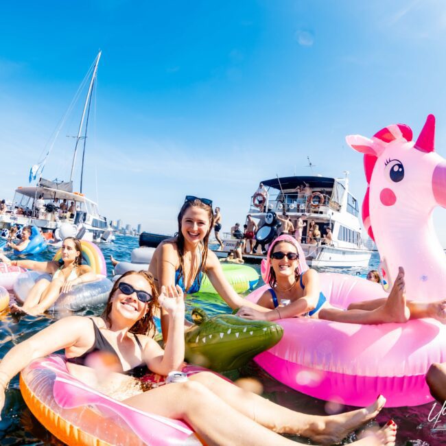 A group of smiling people relax on colorful inflatable pool floats, including a large pink unicorn, in the water. Boats are anchored nearby on a clear, sunny day. The scene conveys a fun and festive atmosphere.