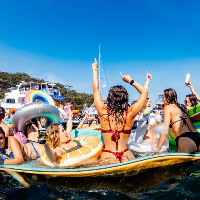 A vibrant scene of people enjoying a yacht party. They are on inflatables in the water, with some holding drinks and dancing. The background features yachts, clear blue sky, and a forested shoreline.
