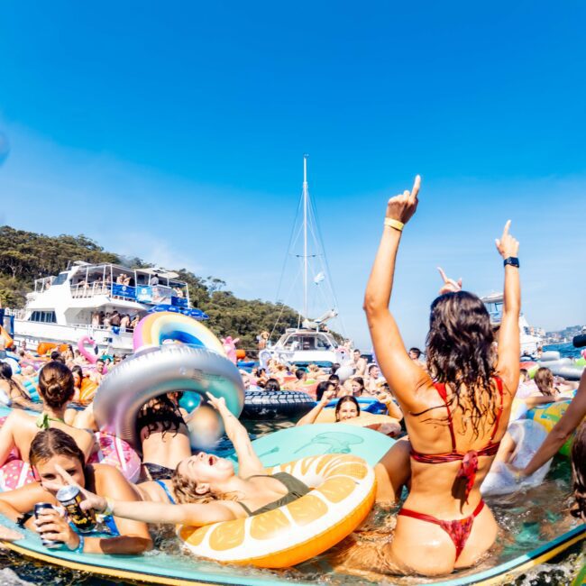 A festive scene of people enjoying a sunny day on the water, floating on colorful inflatable rafts shaped like fruit. Boats are in the background, and the atmosphere is lively and cheerful, with music likely playing.