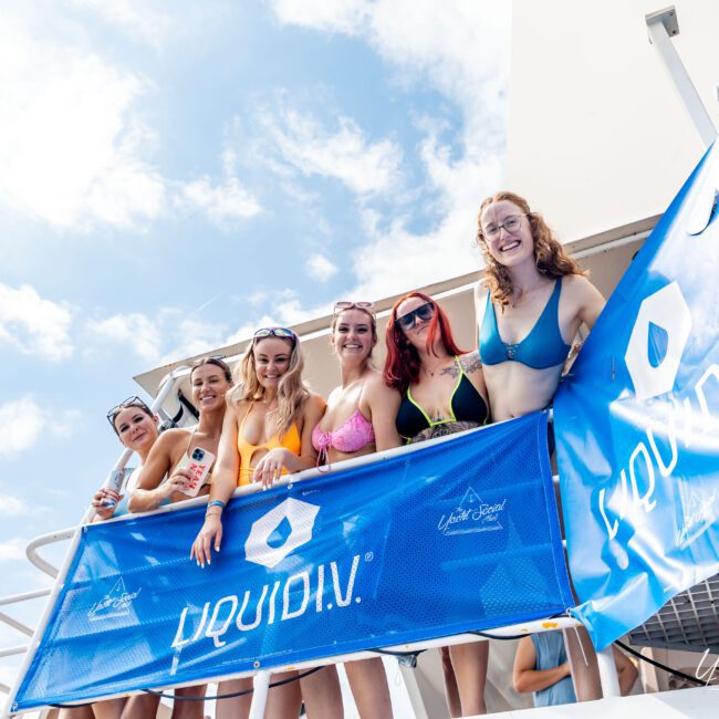 A group of seven women in swimwear stand on a boat's deck behind a blue Liquid I.V. banner. They are smiling and posing for the camera under a partly cloudy sky.