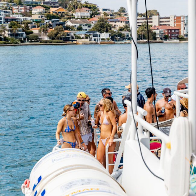 A group of people socializing and sunbathing on a boat, anchored near a coastal city with houses visible on a hillside. Some are wearing swimsuits and chatting, while others stand near the boat's railing. It is a sunny day.