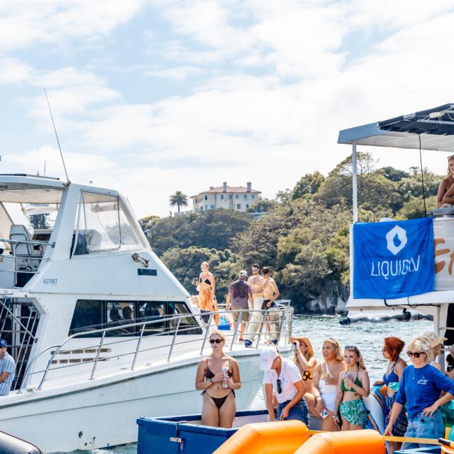 People in swimwear are enjoying a boat party with inflatable slides on the water. Two boats are docked side by side, surrounded by greenery and a hillside house in the background. The scene is sunny with blue skies.