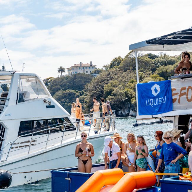 A lively gathering on boats with people partying and relaxing by the water. One boat has a branded canopy, and inflatables are visible in the water. Trees and a building on a hill are in the background under a partly cloudy sky.