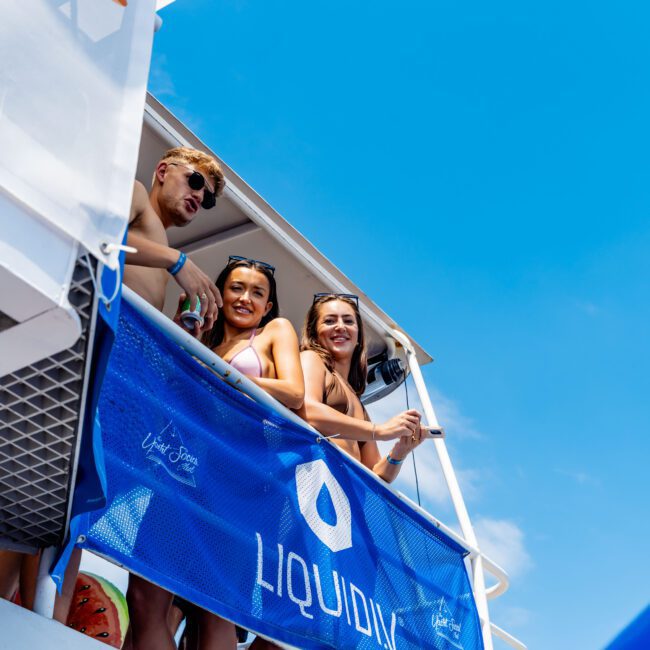 Three people are smiling and relaxing on a boat deck, leaning on a blue banner with the text "LIQUID." The sky is clear and blue, and there's a logo in the bottom right corner with the text "The Yacht Social Club.